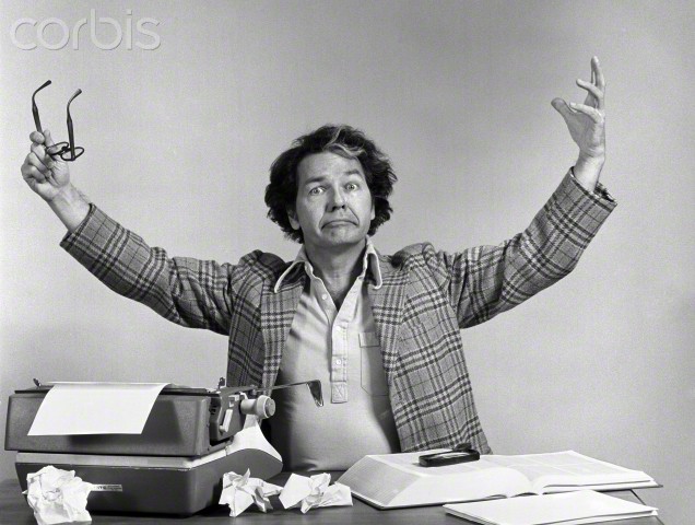 23 Sep 1976 --- 1970s MALE WRITER AT DESK TYPEWRITER CRUMPLED PAPERS ARMS UP IN AIR EXASPERATED EXPRESSION INDOOR LOOKING AT CAMERA --- Image by © H. ARMSTRONG ROBERTS/Corbis